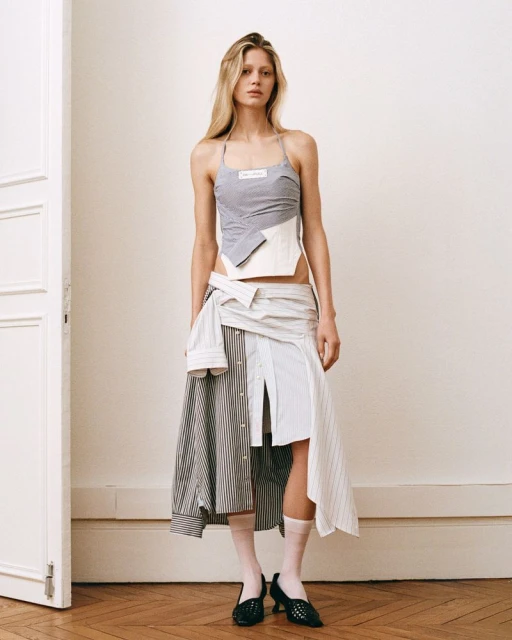 A fashionable adult female in a unique grey halter top and mixed-fabric skirt stands against an off-white wall with a partially visible white door.