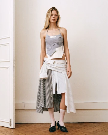 A fashionable adult female in a unique grey halter top and mixed-fabric skirt stands against an off-white wall with a partially visible white door.