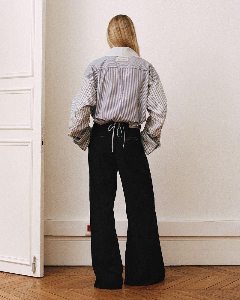 Blonde woman wearing a striped oversized button-down shirt and black wide-leg pants standing in a room with a wooden floor.