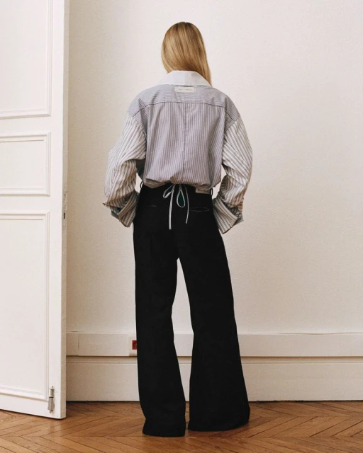 Blonde woman wearing a striped oversized button-down shirt and black wide-leg pants standing in a room with a wooden floor.