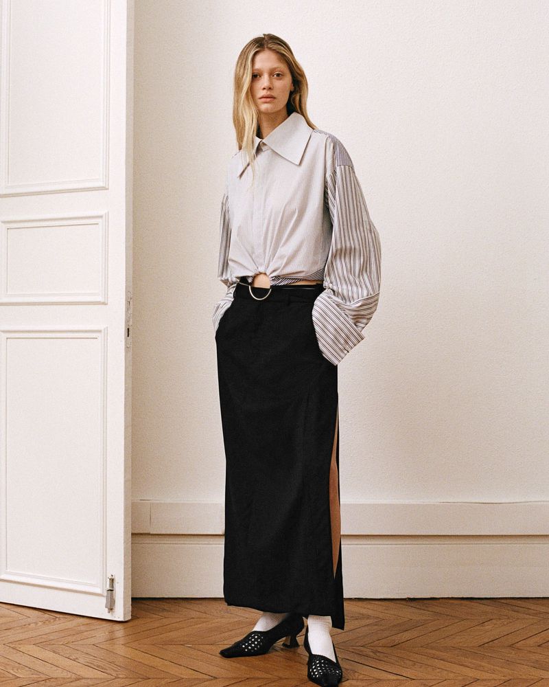 Fashionable adult woman poses in a stylish white and striped collared shirt and black maxi skirt with leg slit, inside an indoor setting.