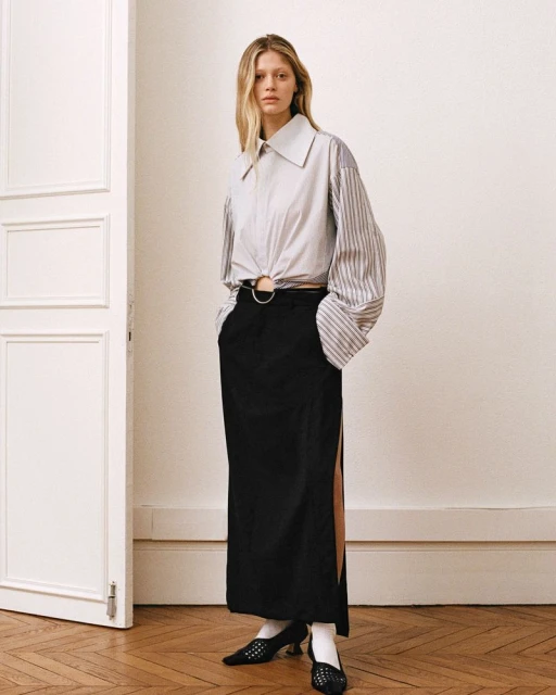 Fashionable adult woman poses in a stylish white and striped collared shirt and black maxi skirt with leg slit, inside an indoor setting.