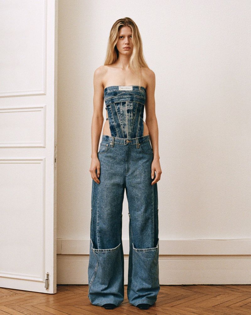 Full shot of a model in a blue denim strapless top and wide-legged jeans, posing in front of a white wall with wooden floors.