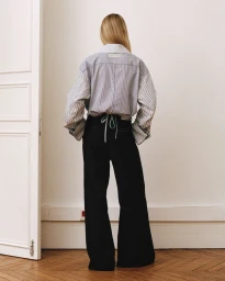 Model seen from behind wearing a striped button-down shirt and black trousers against an off-white wall