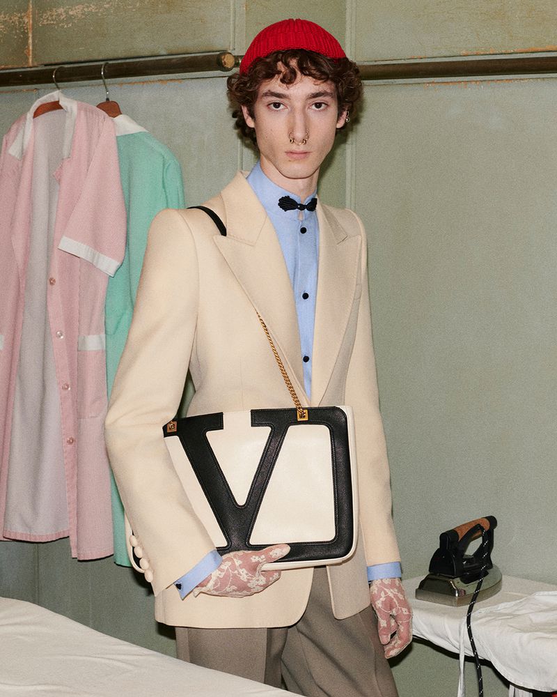 A stylish young man in a beige suit, bowler, and lace gloves holds a Valentino bag on a vintage ironing board in an indoor scene.
