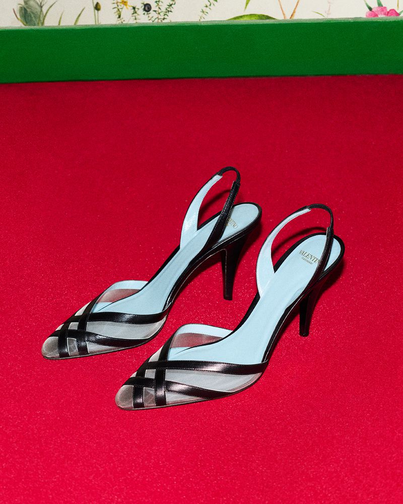 Elegant pair of black heeled pumps with a sheer net and black leather strap design on a vibrant red surface, sophisticated footwear.