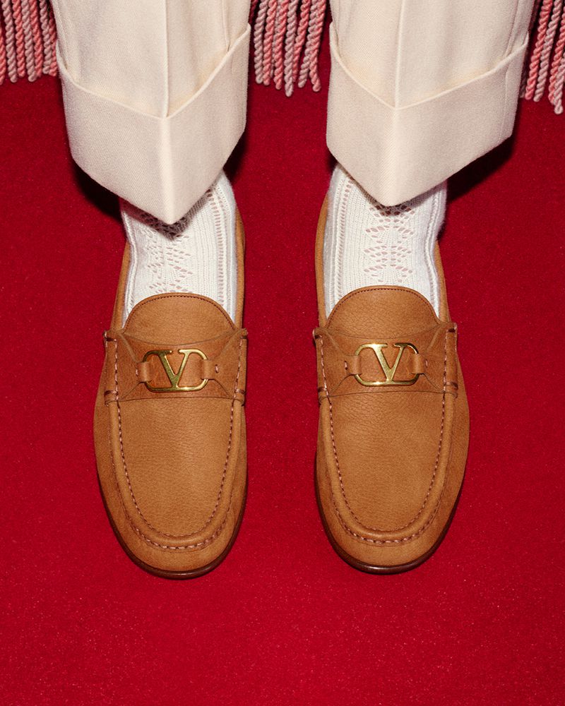 Tan leather Valentino loafers with gold logo hardware are paired with textured white socks and cream trousers on a red carpet.