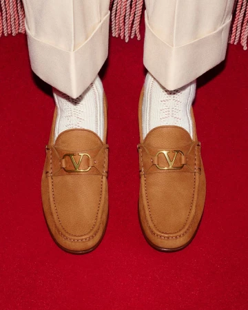 Tan leather Valentino loafers with gold logo hardware are paired with textured white socks and cream trousers on a red carpet.