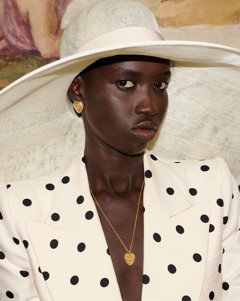 A portrait of a Black woman wearing a large-brimmed hat and a polka-dot blazer, along with a gold necklace and earrings. 