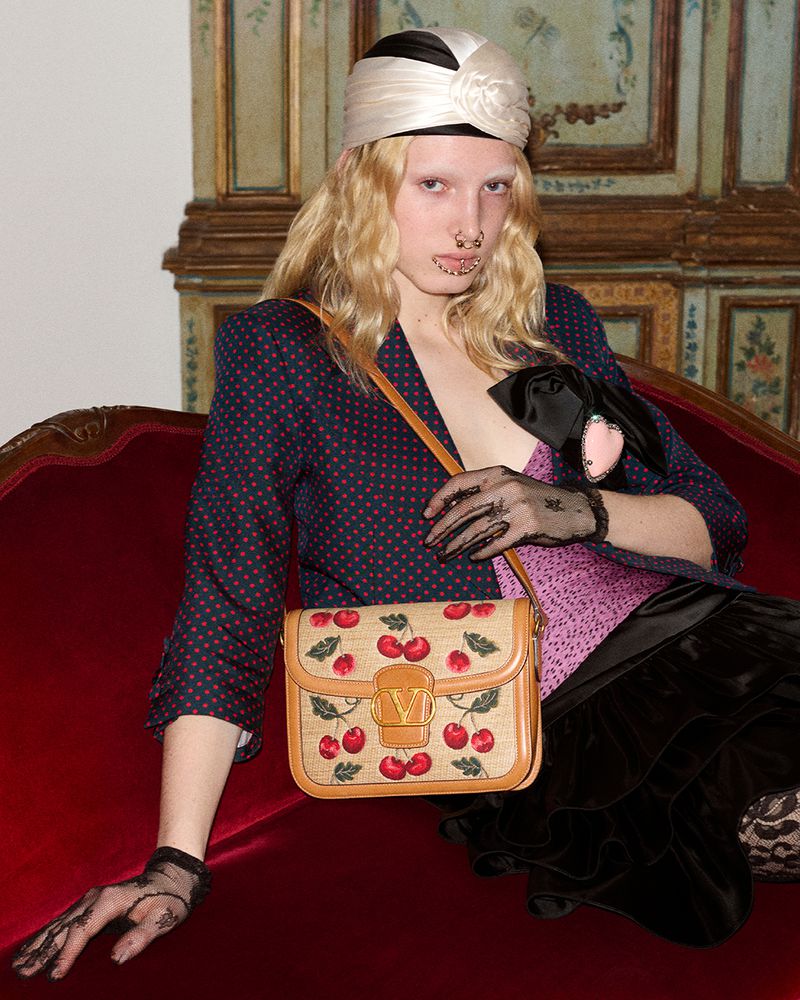 A woman poses on a red velvet couch wearing a polka dot blazer, turban, and holding a handbag with cherry motifs in a fashion studio.