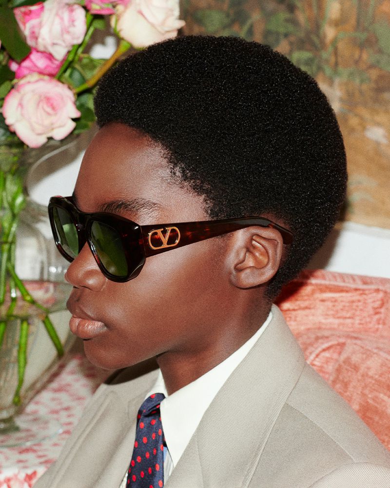 Young adult wearing sunglasses with a beige suit and patterned tie, posing in a room with a floral arrangement on a table.