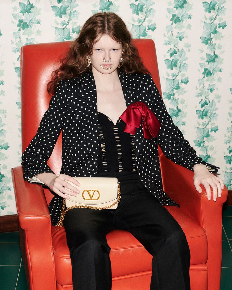 Fashion shot of a model in a polka dot blazer sitting in an orange chair, holding a beige Valentino bag with gold chain strap.