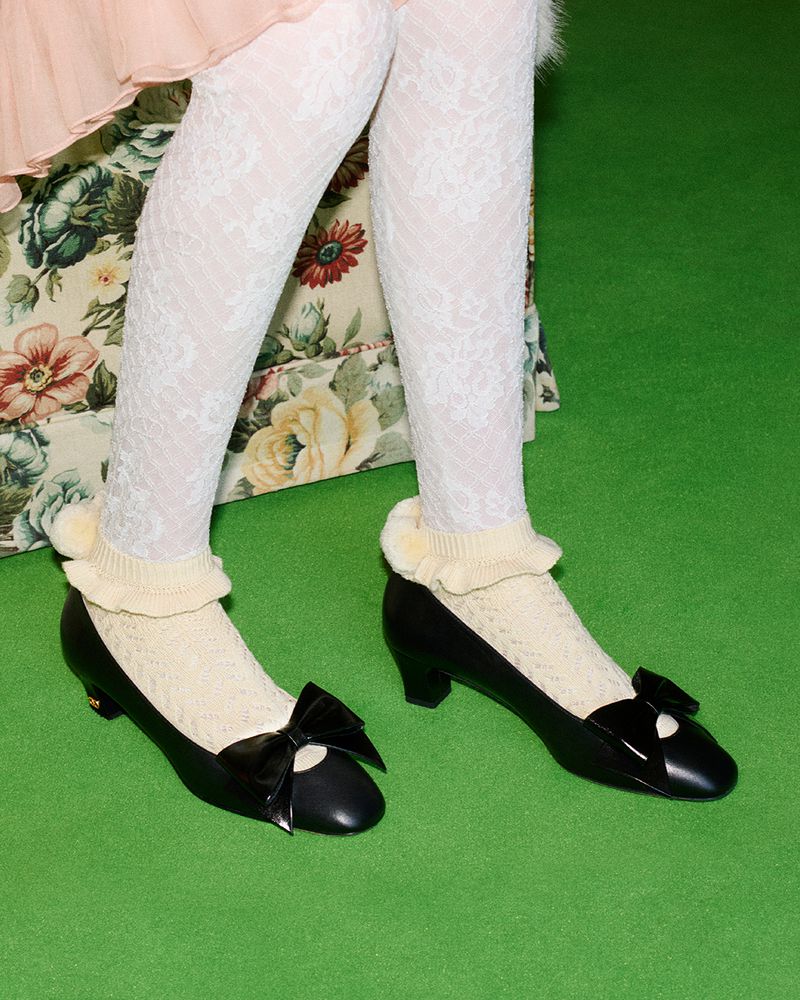 Close-up of a person wearing white lace tights and black leather flats with bows, standing on a bright green surface. Floral pattern chair.