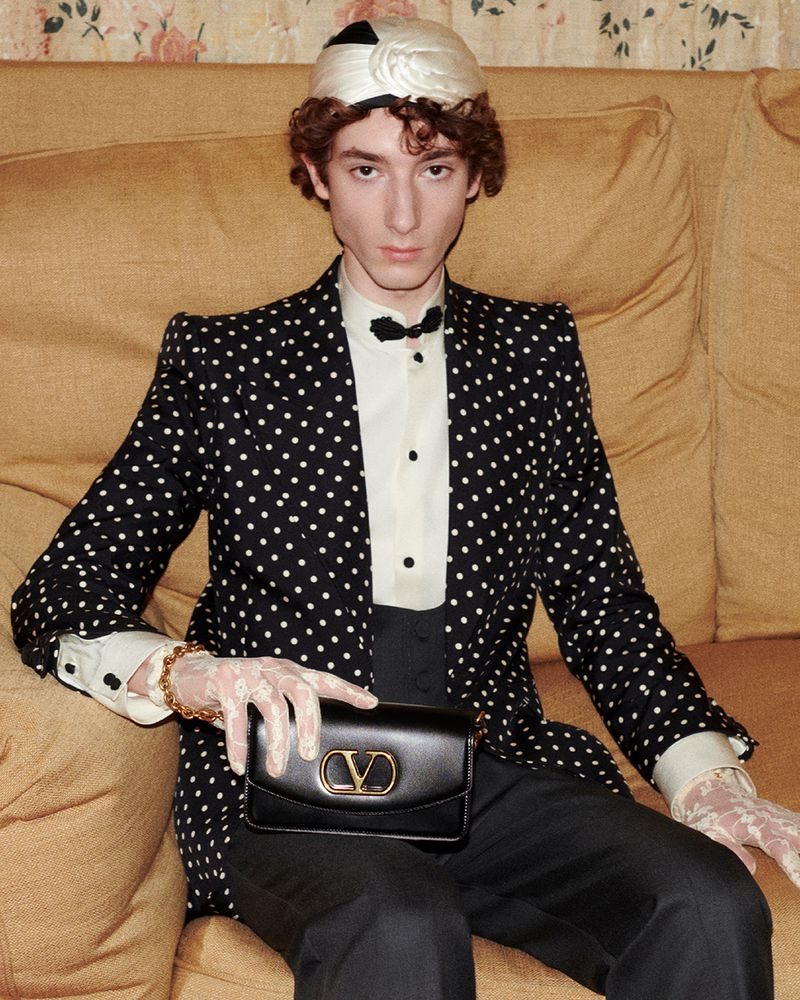 A person in a polka-dot blazer, bow tie, and white cap poses with a black leather purse while sitting on a tan couch.