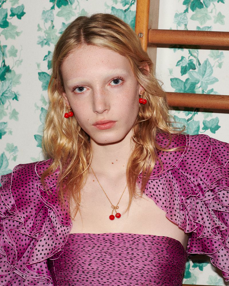 A fair-skinned young woman models cherry earrings and necklace in a pink polka dot ruffled outfit against a floral wallpaper.