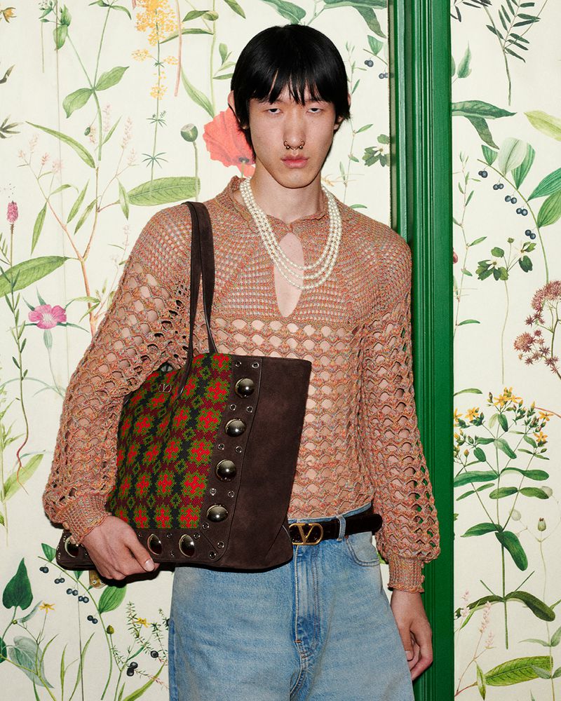 An Asian male fashion model wearing a tan sweater, jeans, and pearl necklace, carrying a patterned tote bag in a studio setting.