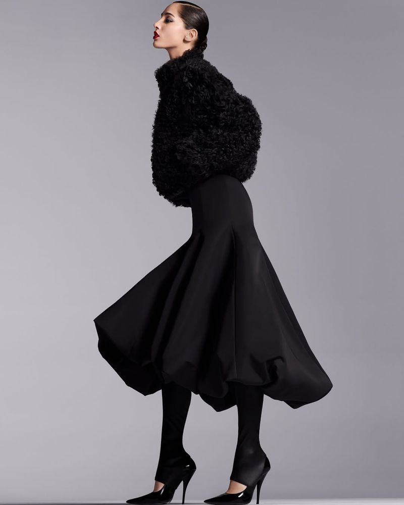 A stylish woman in a black, puffy jacket and full skirt poses in profile against a grey backdrop, showcasing fashion and elegance.