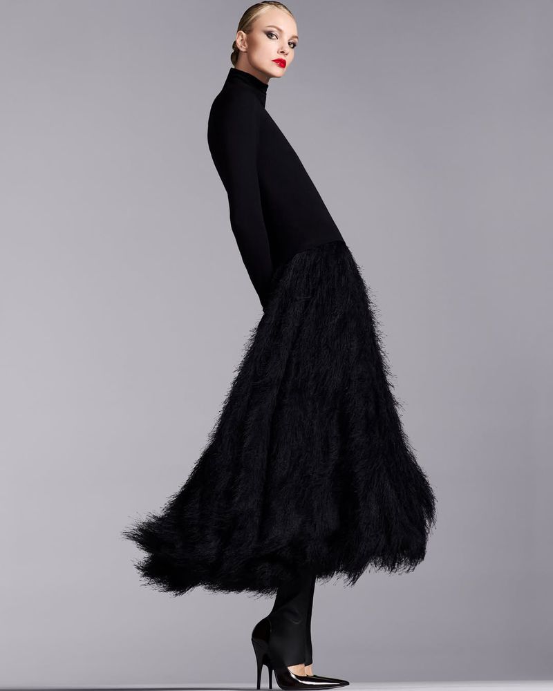 Woman in long-sleeved black dress with feathered skirt, wearing black high heels in a studio with gray backdrop. Fashion photography.