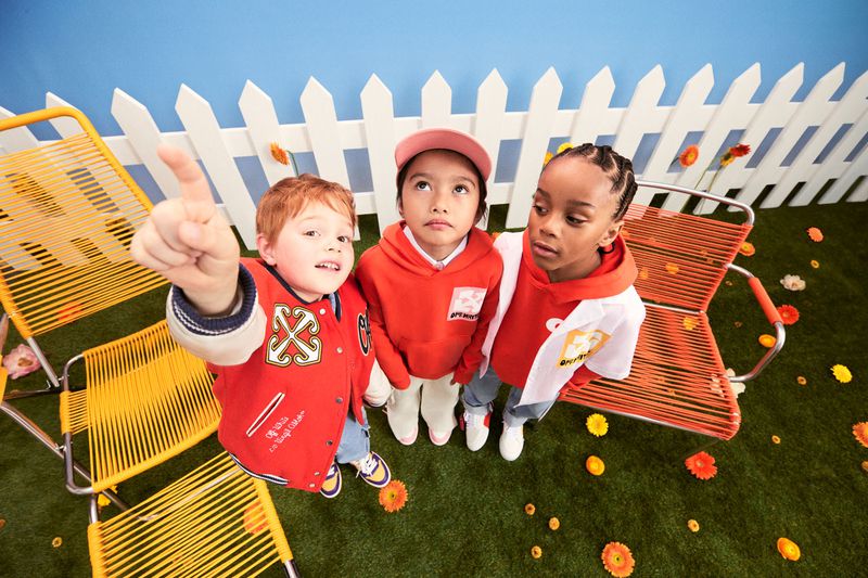 Three young children stand in an artificial yard setting with chairs and flowers, wearing colorful outfits, looking up at something out of frame.