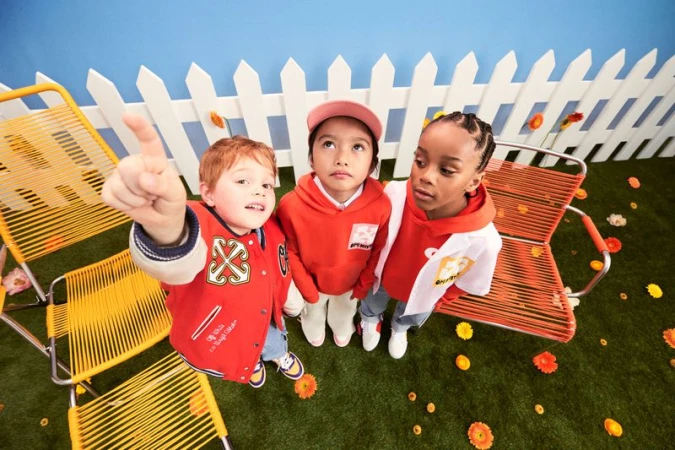 Three young children stand in an artificial yard setting with chairs and flowers, wearing colorful outfits, looking up at something out of frame.