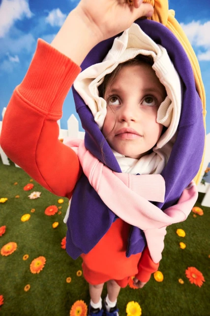 Child in multi-colored hoodies in a field of flowers. The sky is blue with clouds and the camera angle is low and looking up.