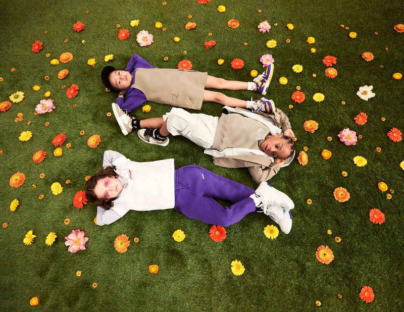Three children lie on a green grass-like surface dotted with orange, yellow, and pink flowers, viewed from above, capturing a playful, relaxed moment.