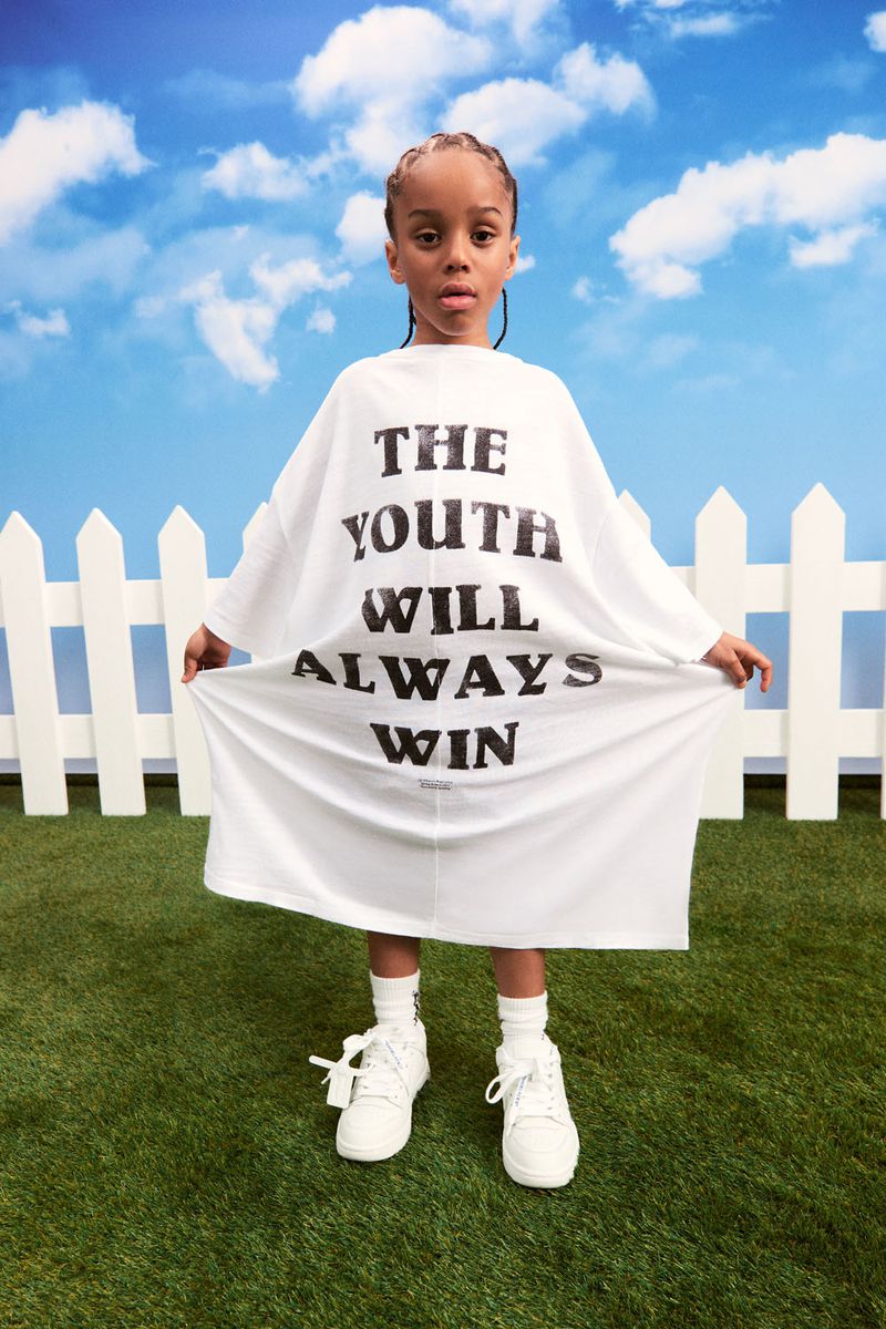 Child in oversized white t-shirt with 'THE YOUTH WILL ALWAYS WIN' text stands on green grass against a blue sky backdrop.