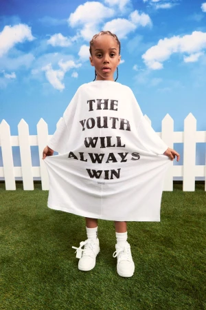 Child in oversized white t-shirt with 'THE YOUTH WILL ALWAYS WIN' text stands on green grass against a blue sky backdrop.