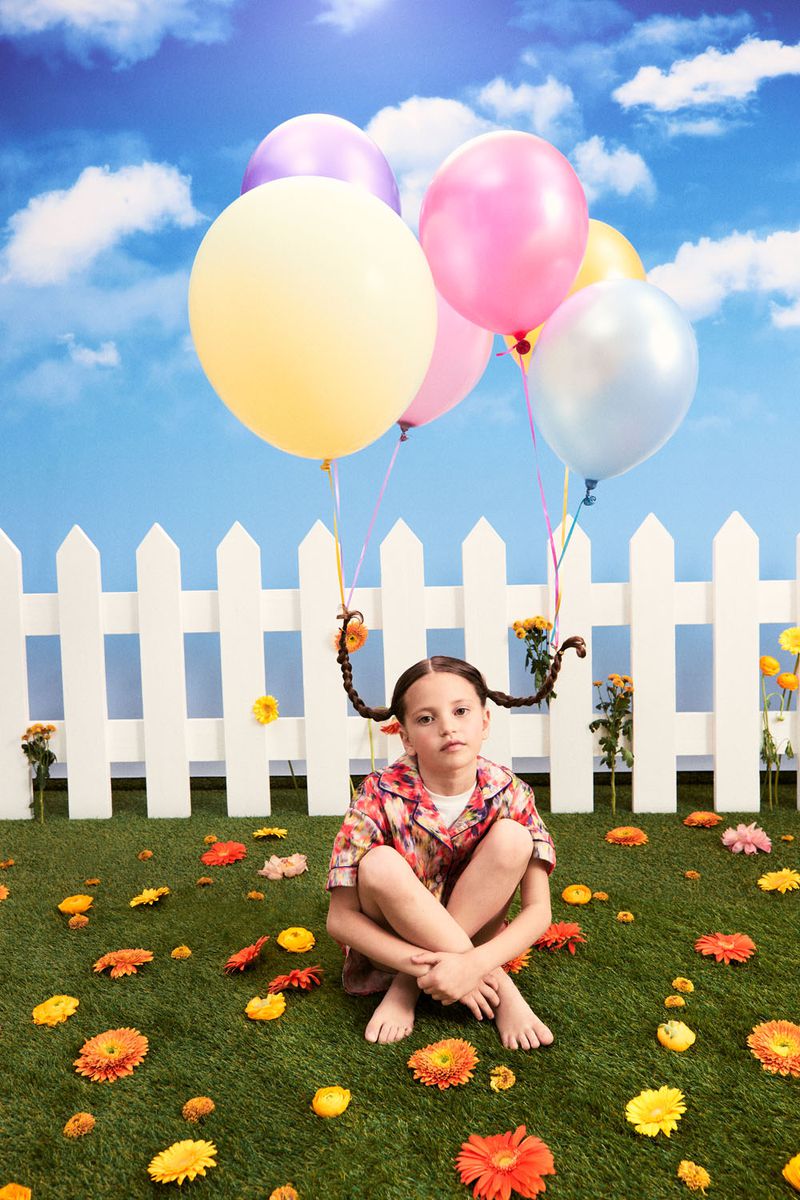Young girl sits with balloons on grassy lawn with flowers in front of white picket fence under a blue sky with white clouds.