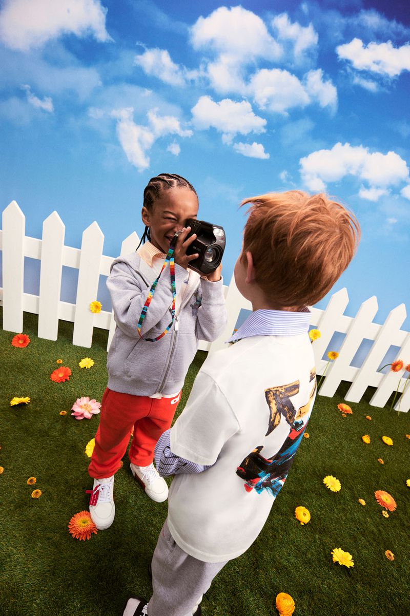 Two children playing outside on a vibrant green lawn with colorful flowers. One is taking a picture with an old-fashioned camera.
