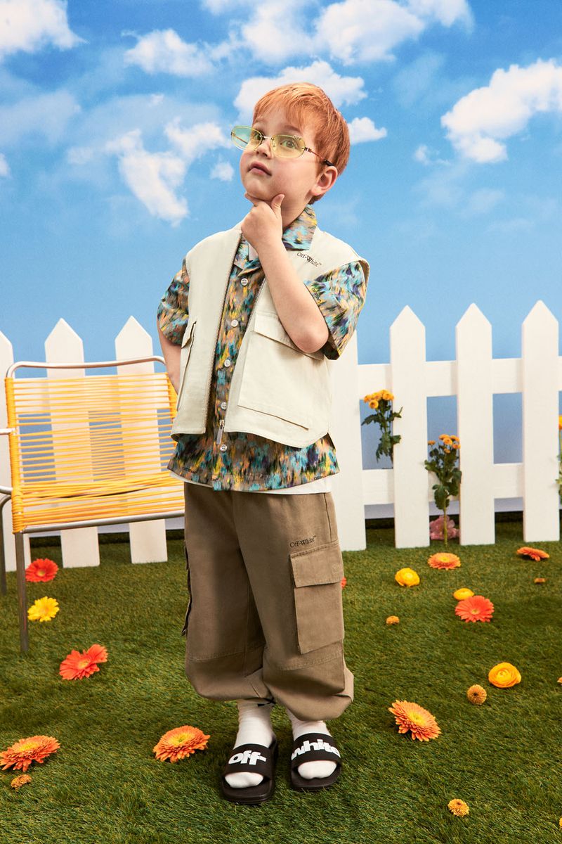 Young boy with red hair poses outdoors wearing sunglasses, a vest, printed shirt, cargo pants, and slides against a sky backdrop.