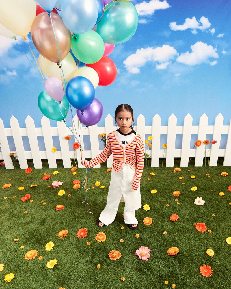 Young girl in a striped sweater and white pants standing in a field holding colorful balloons, with a white picket fence.