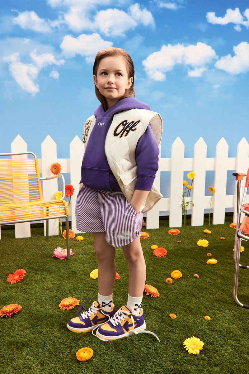 A child model poses outdoors wearing purple hoodie, shorts, sneakers, and a quilted vest in a playful outdoor setting with flowers and blue sky.