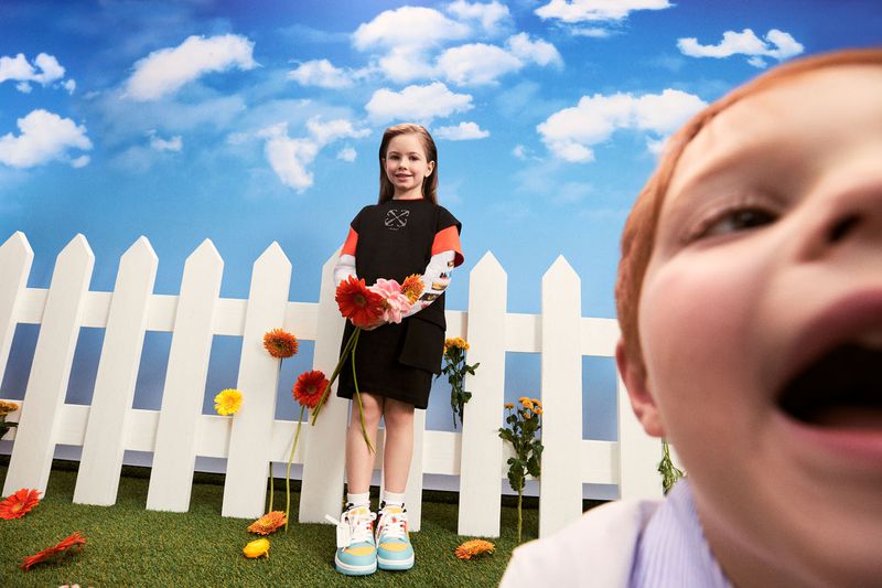 A young girl in a black dress holds a bouquet of flowers in front of a white picket fence with a blue sky and a child's face to the right.