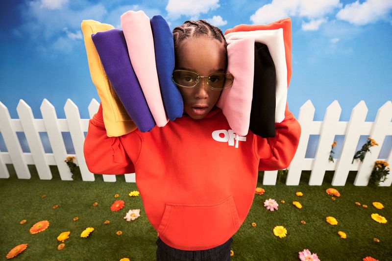 Child model wearing an orange hoodie and glasses outdoors, carrying folded colorful hoodies. Fashion editorial on faux grass with white picket fence.