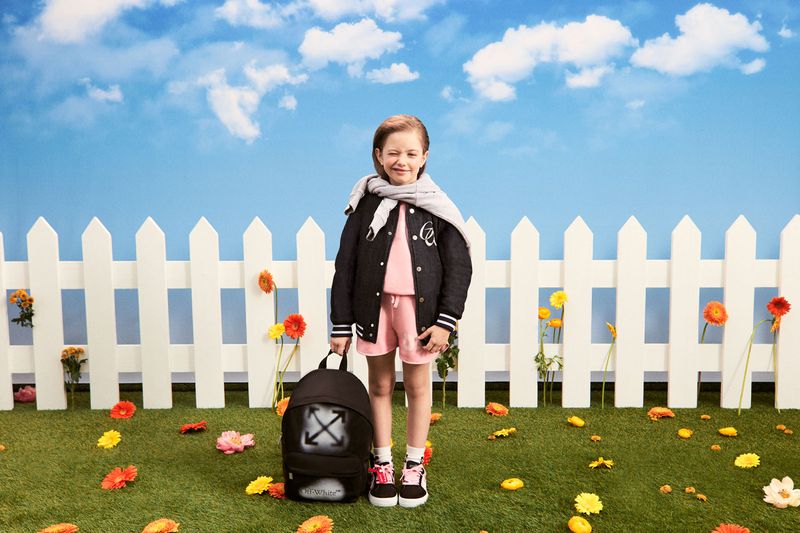 Young girl poses in front of a white picket fence on green grass with a black backpack, wearing a black jacket and scarf.