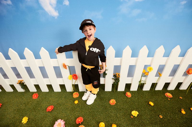A young boy in black athletic wear stands in front of a white picket fence on artificial grass with scattered orange flowers.