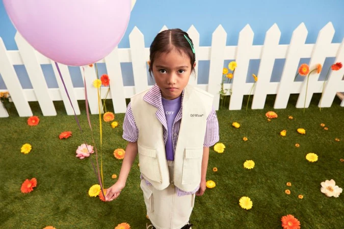 Young girl in vest and skirt holds a pink balloon outdoors against a white picket fence and artificial grass strewn with flowers.