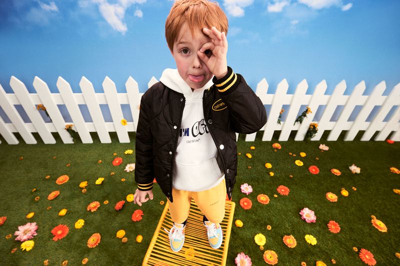 Young red-haired child poses on yellow grate, making an 'ok' sign near a white picket fence with artificial grass and scattered flowers.