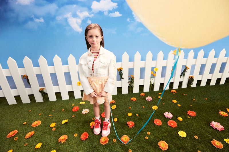Young girl in a white jacket and skirt standing on grass scattered with orange flowers, holding a yellow balloon against a sky background.