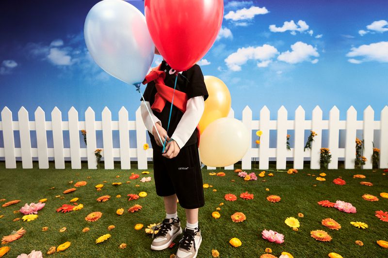 Child stands in a grassy field in front of a white picket fence, face obscured by red, yellow, and white balloons against a sky backdrop.
