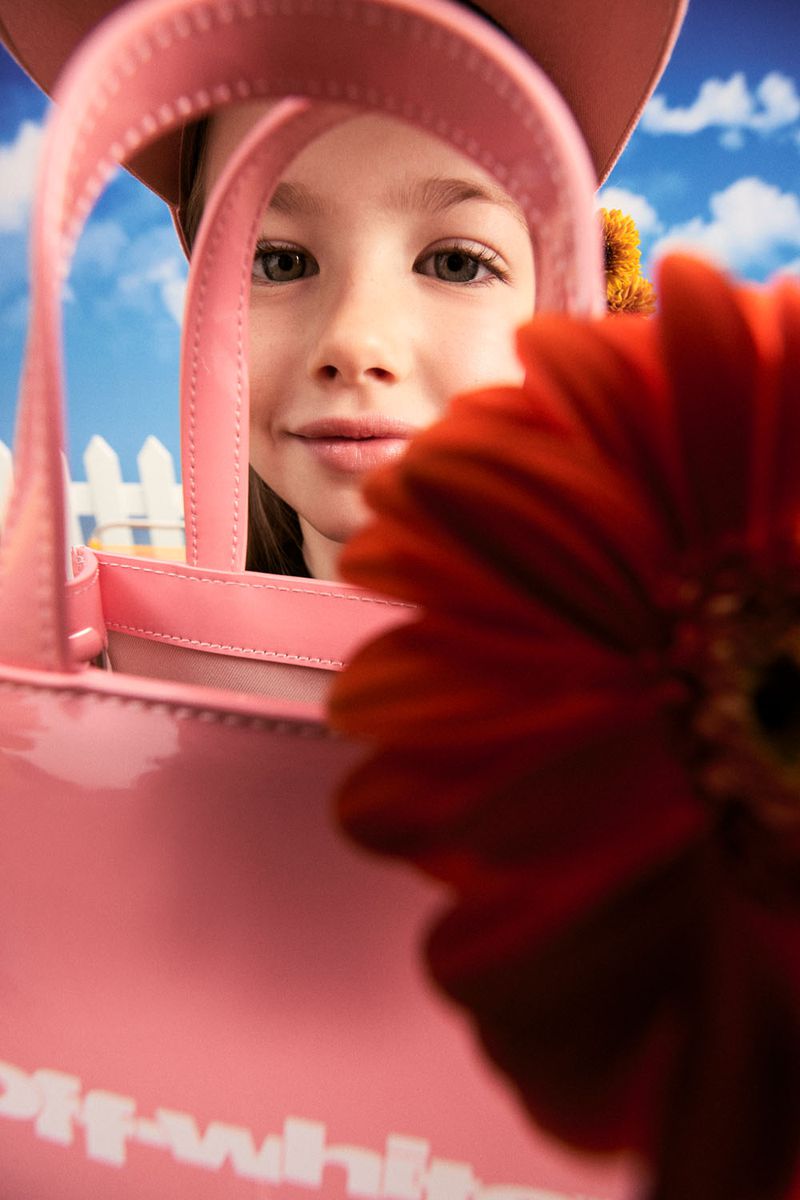 A child model displays a pink 'Off-White' handbag indoors, with a blurred red flower and blue sky backdrop, highlighting youth fashion.