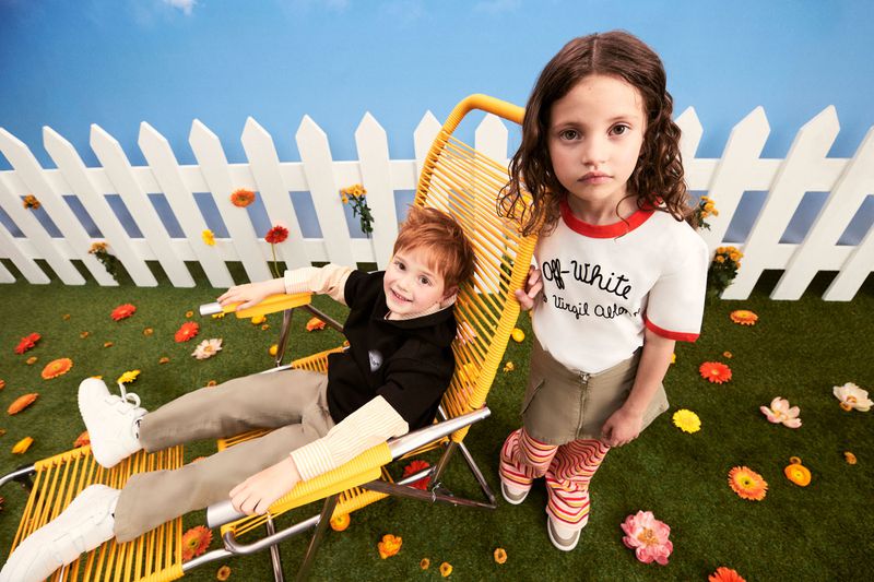 Two children pose on a lawn with scattered flowers against a picket fence and blue sky, one child lounging in a yellow lawn chair.