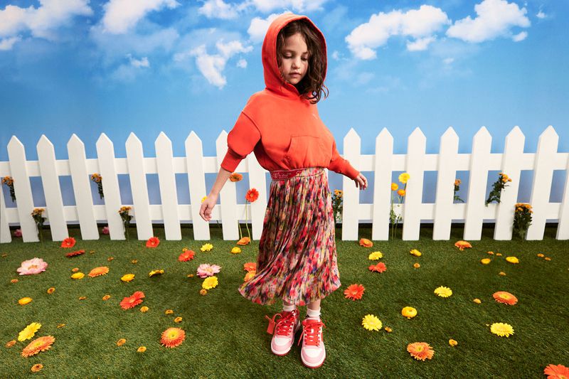 Child in orange hoodie and floral skirt stands in a sunny, flowered field by a white picket fence against a blue sky with clouds.