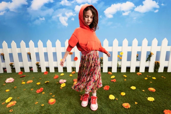 Child in orange hoodie and floral skirt stands in a sunny, flowered field by a white picket fence against a blue sky with clouds.