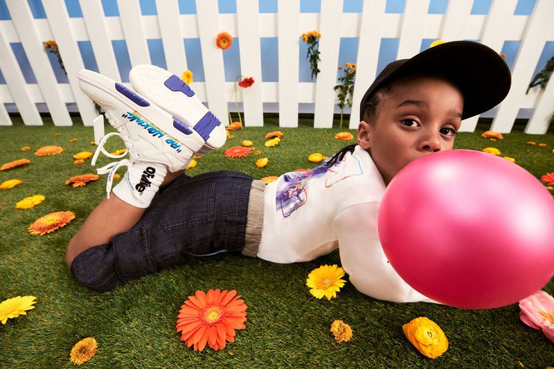A young child lies on artificial turf surrounded by flowers, blowing up a large pink balloon, wearing a black cap, jeans, and sneakers.