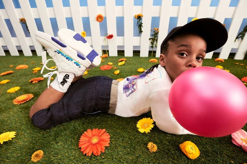 A young child lies on artificial turf surrounded by flowers, blowing up a large pink balloon, wearing a black cap, jeans, and sneakers.