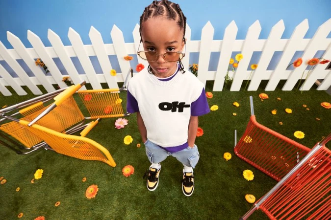 Child models apparel in a whimsical setting with flowers and scattered chairs, captured from an eye-level angle with a white picket fence.