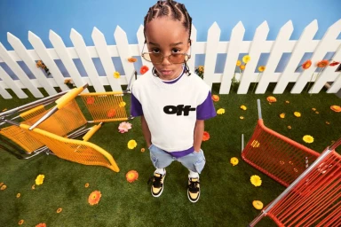 Young child in white tee and sunglasses standing on artificial grass with scattered flowers and upturned chairs.