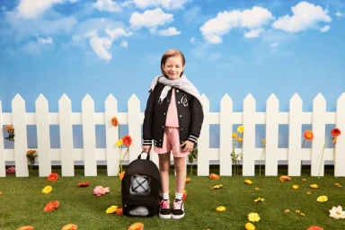 Child model wearing a black varsity jacket and pink shorts standing in front of a white picket fence under a blue sky backdrop.